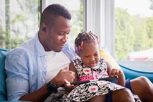 fröhlicher afroamerikanischer vater und tochter spielen im wohnzimmer, süßes kleines mädchen und papa sitzen auf dem sofa und spielen auf dem tablet, glücksfamilienkonzepte foto