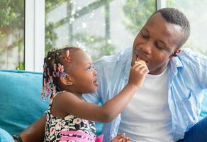 süßes kleines mädchen, das ihren vater sieht, der einen snack isst, fröhlicher afroamerikanischer vater und tochter, die im wohnzimmer spielen, glücksfamilienkonzepte foto