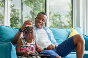 Fröhlicher afroamerikanischer Vater und Tochter spielen im Wohnzimmer, süßes kleines Mädchen sitzt auf dem Sofa und spielt auf dem Tablet foto