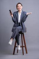 junge asiatische geschäftsfrau, die auf stuhl sitzt und smartphone auf grauem hintergrund verwendet foto