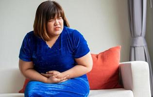 Übergewichtige asiatische Frauen sitzen auf dem Sofa im Wohnzimmer. und Händchenhalten am Bauch wegen Gastritis. Krankheits- und Gesundheitskonzepte foto