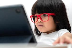 Kleines asiatisches Vorschulmädchen mit roter Brille und Tablet-PC auf weißem Hintergrund, asiatisches Mädchen, das mit einem Videoanruf mit einem Tablet spricht und lernt, Bildungskonzept für Schulkinder. foto