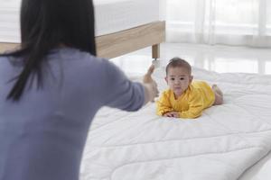 Ein asiatisches Kleinkind, das das Krabbeln mit seiner Mutter übt, die auf der anderen Seite ihres weichen weißen Tuchs im Schlafzimmer sitzt foto