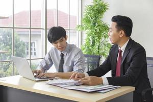 ein asiatischer männlicher vorgesetzter bringt dem neuankömmling das büro auf seinem schreibtisch bei. foto