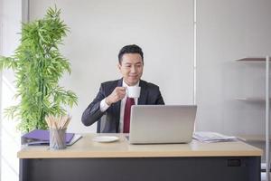 ein asiatischer manager, der in einem büro arbeitet und eine weiße kaffeetasse mit einem strahlenden lächeln hält. foto