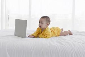 Ein asiatisches Baby spielt auf einem Laptop auf einem sauberen weißen Bett. foto