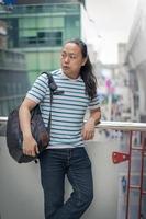 Ein thailändisch-chinesischer Typ mit langen schwarzen Haaren und T-Shirt mit blauer Linie denkt nach und hält einen Rucksack auf seiner Schulter in der Nähe des Sky Train. foto