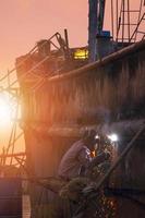 Seitenansicht des Schweißers in Schutzkleidung auf der Plattform schweißt die Metallwand des alten Fischereifahrzeugs im Werftbereich bei Sonnenuntergang im vertikalen Rahmen foto
