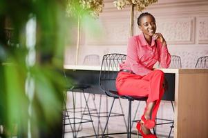 hübsche Zöpfe Business Afroamerikanerin hell herrisch Person freundliche Kleidung Büro rotes Hemd und Hose, auf Stuhl sitzend. foto