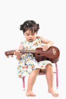 asiatisches süßes Baby, das Ukulele spielt, sitzt isoliert auf weißem Hintergrund. Baby-Musik-Konzept. foto