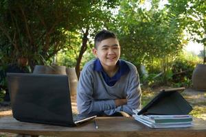 Junge, der morgens in einem ländlichen Haus auf die Kamera schaut, die online auf einem Holztisch mit Laptop und Tablet studiert. Konzept Online-Bildung auf dem Land und Arbeit von zu Hause aus foto