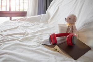 Ein roter Kopfhörer wird morgens auf ein Buch in der Nähe eines rosa Teddybären auf dem Bett gelegt, um sich bequem auszuruhen. ein kopfhörer und ein buch ruhen auf der weißen bettwäsche im schlafzimmer.