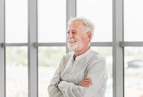 gutaussehender älterer kaukasischer alter mann, der mit verschränkten armen lächelt, glücklicher älterer mann, der in der nähe des fensters steht foto