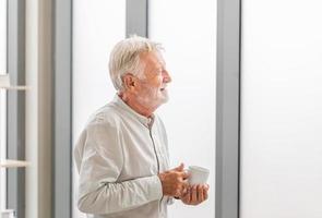 älterer Mann im neuen Zuhause während einer Kaffeepause, älterer Mann, der mit Tassen Kaffee in der Nähe des Fensters steht foto