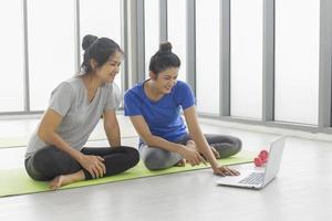 zwei asiatinnen mittleren alters schauen sich an, wie man von ihren laptops aus im fitnessstudio grundlegendes yoga macht. foto