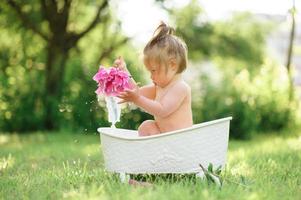 glückliches Kleinkind nimmt ein Milchbad mit Blütenblättern. kleines Mädchen in einem Milchbad auf grünem Hintergrund. Blumensträuße aus rosa Pfingstrosen. baby baden. Hygiene und Pflege für Kleinkinder. foto