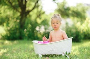 glückliches Kleinkind nimmt ein Milchbad mit Blütenblättern. kleines Mädchen in einem Milchbad auf grünem Hintergrund. Blumensträuße aus rosa Pfingstrosen. baby baden. Hygiene und Pflege für Kleinkinder. foto