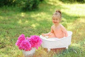 glückliches Kleinkind nimmt ein Milchbad mit Blütenblättern. kleines Mädchen in einem Milchbad auf grünem Hintergrund. Blumensträuße aus rosa Pfingstrosen. baby baden. Hygiene und Pflege für Kleinkinder. foto