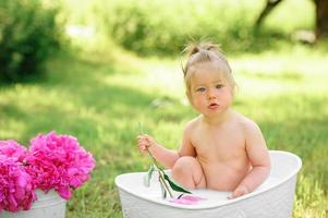 glückliches Kleinkind nimmt ein Milchbad mit Blütenblättern. kleines Mädchen in einem Milchbad auf grünem Hintergrund. Blumensträuße aus rosa Pfingstrosen. baby baden. Hygiene und Pflege für Kleinkinder. foto