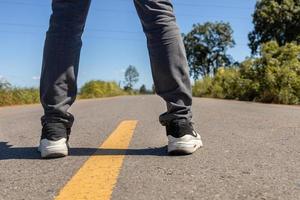 Mannfüße stehen auf Asphaltstraße mit gelben Markierungslinien. Mann mit Turnschuhen und Jeans. foto