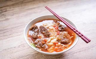 Rindfleisch-Nudel-Ramen-Mahlzeit mit Tomatensauce-Brühe in Schüssel auf hellem Holztisch, berühmtes Essen im chinesischen Stil in Taiwan, Nahaufnahme, Draufsicht, Kopierraum foto