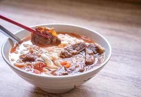 Rindfleisch-Nudel-Ramen-Mahlzeit mit Tomatensauce-Brühe in Schüssel auf hellem Holztisch, berühmtes Essen im chinesischen Stil in Taiwan, Nahaufnahme, Draufsicht, Kopierraum foto