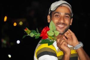 junger indischer mann lächelt glücklich und hält rosenblume in der nacht foto
