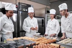 Multirassisches professionelles Gourmet-Team, vier Köche in weißen Kochuniformen und Schürzen kneten Teig und Mehl, bereiten Brot und Backwaren zu und backen im Ofen in der Restaurantküche aus Edelstahl. foto