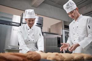 zwei professionelle asiatische männliche köche in weißen kochuniformen und schürzen kneten teig und eier, bereiten brot und frische backwaren zu und backen im ofen in der edelstahlküche des restaurants. foto