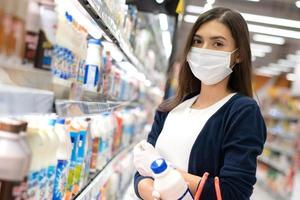 schöne frau mit medizinischer gesichtsmaske und gummihandschuh mit einkaufswagen, der täglich milch im produktregal abholt. einkaufen im supermarkt in einem neuen normalen lebensstilkonzept während des coronavirus. foto