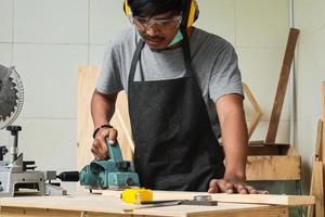 ein junger männlicher Zimmermann, der mit Elektrohobel auf Holzplanken in seiner Werkstatt arbeitet und Sicherheitsausrüstung trägt foto