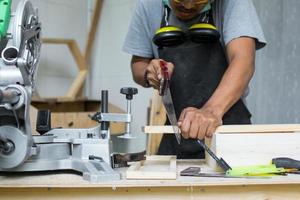 ein junger männlicher Zimmermann, der das Holz mit einer Handsäge auf seinem Werkstatttisch mit Sicherheitsausrüstung schneidet foto