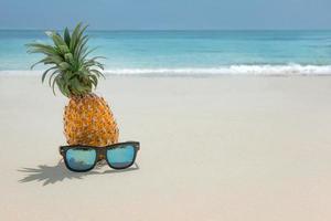 ananasfrucht in sonnenbrille auf sand gegen türkisfarbenes karibisches meerwasser. tropisches sommerferienkonzept