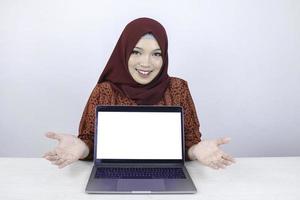 junge asiatische islamfrau lächelt und zeigt auf weißem leerem bildschirm auf dem laptop zur kamera. foto