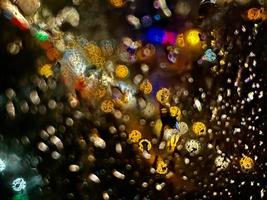 Schuss durch das Glas im Regen mit Straßenlaternen und Autoscheinwerfern im Hintergrund. verschwommener Bokeh-Glitzer auf hellem Hintergrund foto