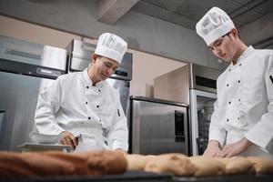 zwei professionelle asiatische männliche köche in weißen kochuniformen und schürzen kneten teig und eier, bereiten brot und frische backwaren zu und backen im ofen in der edelstahlküche des restaurants. foto