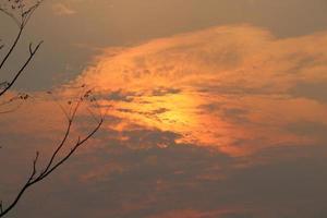 Sonne hinter Wolken und trockenen Zweigen von Bäumen, orangefarbenem und grauem Himmelshintergrund. foto