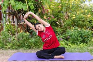 Fröhliche junge asiatische Frau, die Dehnungsübungen auf einer Yogamatte im Freien auf einem natürlichen grünen Hintergrund wärmt. foto