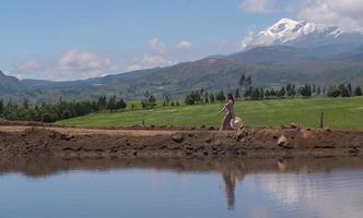 schöne hispanische frau geht mit einem kleid und einem hut auf einer unbefestigten straße mit dem cayambe-vulkan im hintergrund während eines sonnigen morgens und eines blauen himmels foto