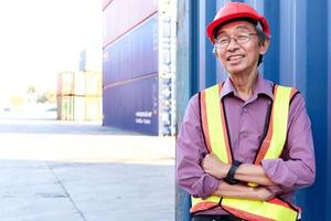 porträt eines glücklich lächelnden älteren asiatischen arbeiteringenieurs mit sicherheitsweste und helm, der mit verschränkten armen mit blauen containern als hintergrund auf dem hof der logistischen frachtcontainer steht. foto