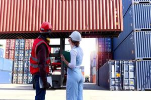 hinter zwei arbeitern tragen sicherheitsweste und helm, die auf dem hof der logistischen schiffsfrachtcontainer diskutieren. afrikanisch-amerikanischer ingenieurmann spricht mit schöner junger chefin mit blonden haaren am arbeitsplatz. foto