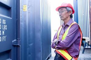 Porträt eines ernsthaften älteren asiatischen Arbeiteringenieurs mit Sicherheitsweste und Helm, der mit verschränkten Armen mit blauen Containern als Hintergrund auf dem Hof der Logistikschifffahrt steht. foto