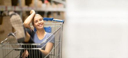 Fröhliche Kundin mit Lächeln und Blick nach oben, während sie im Einkaufswagen im Baumarkt sitzt foto