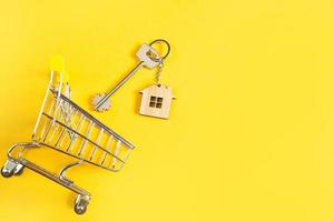 Schlüsselanhänger in Form eines Holzhauses mit Schlüssel auf gelbem Hintergrund und Einkaufswagen. Hypothek, Miete und Kauf von Immobilien, Frühjahrs- und Sommerangebot für den Bau. Platz kopieren foto
