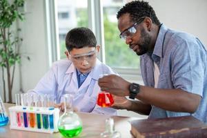 kleiner junge mischt chemikalien in bechern. begeisterter lehrer erklärt kindern chemie, chemiestudent zeigt dem naturwissenschaftsunterricht ein neues experiment foto