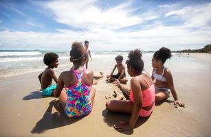 Kinder, die am Strand auf Sand spielen, eine Gruppe von Kindern, die im Sommer am Strand Händchen halten, Rückansicht gegen Meer und blauen Himmel