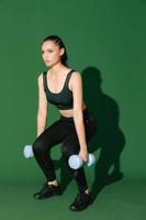 schöne starke fröhliche junge asiatische sportfrau macht übungen mit hantel isoliert auf grünem hintergrund. Fitness-Mädchen in Sportbekleidung, die drinnen posiert. Bewegung, Kraft und Motivation.