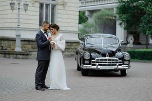 glückliches luxus-hochzeitspaar, das sich im herbst in der nähe von retro mit blumenstraußauto küsst und umarmt foto