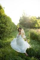 Schwangere Frau mit Hut, die in einem Kleid auf einem Hintergrund von grünen Bäumen posiert. foto