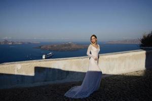 schöne schlanke blonde mädchenfrau topmodel braut mit langen haaren und beinen im weißen brautballkleid mit langer schleppe und volants steht auf einer malerischen insel santorini in griechenland foto